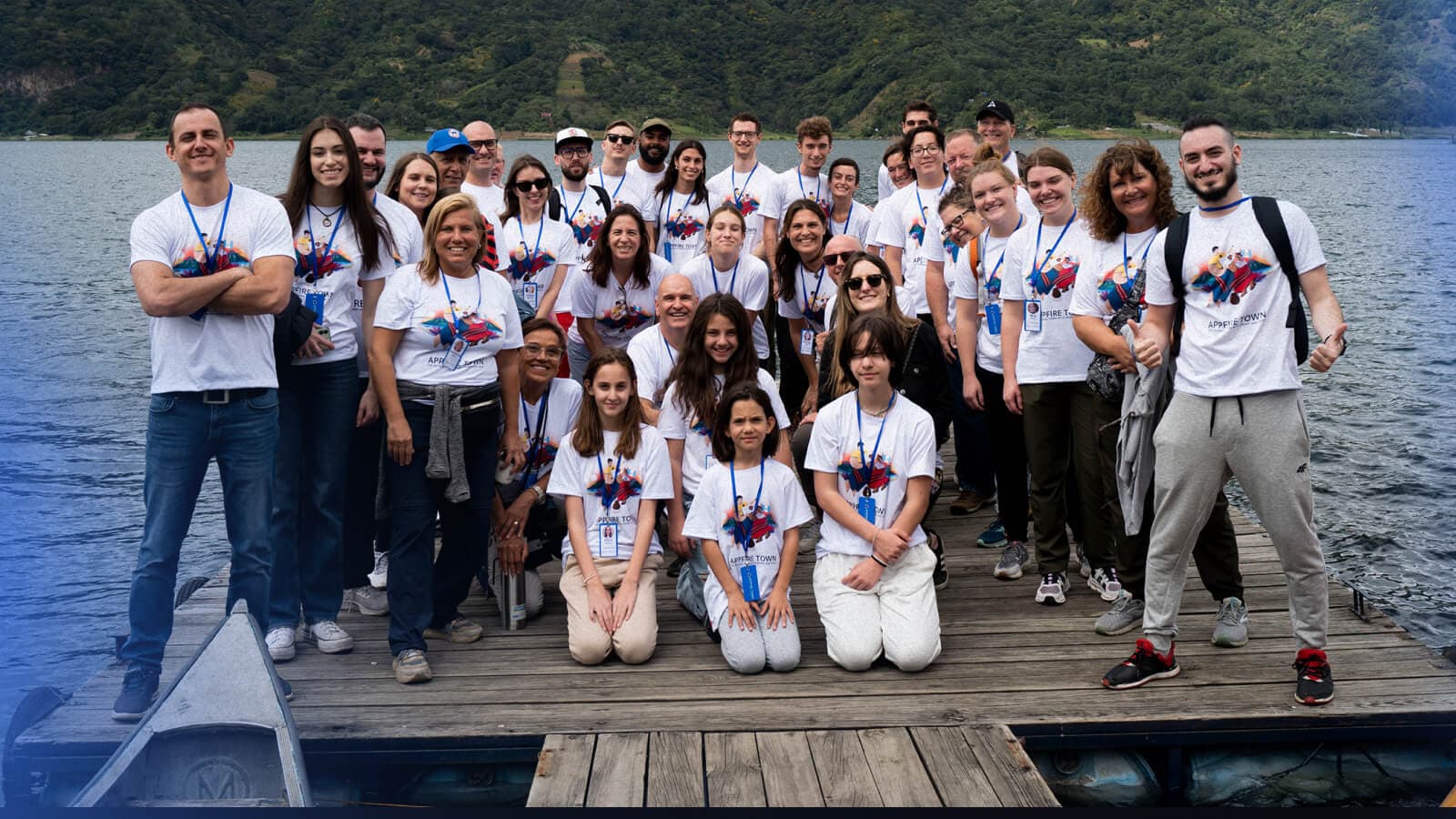 Large group of people gathered on a wooden dock, smiling and posing for a group photo near a lake with mountains in the background.