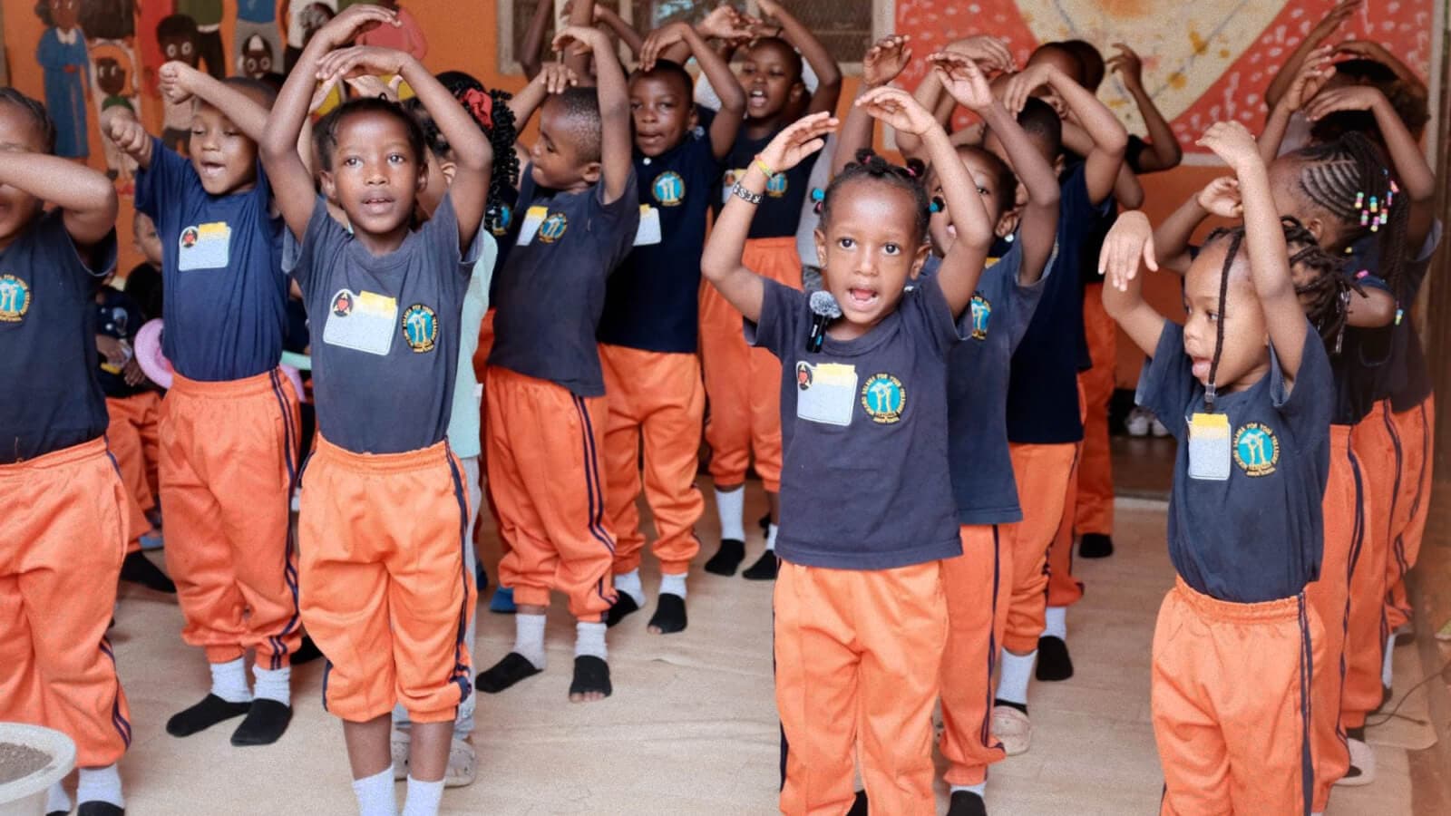 Group of children in orange pants and matching navy shirts standing with arms raised, smiling in a classroom or activity setting.