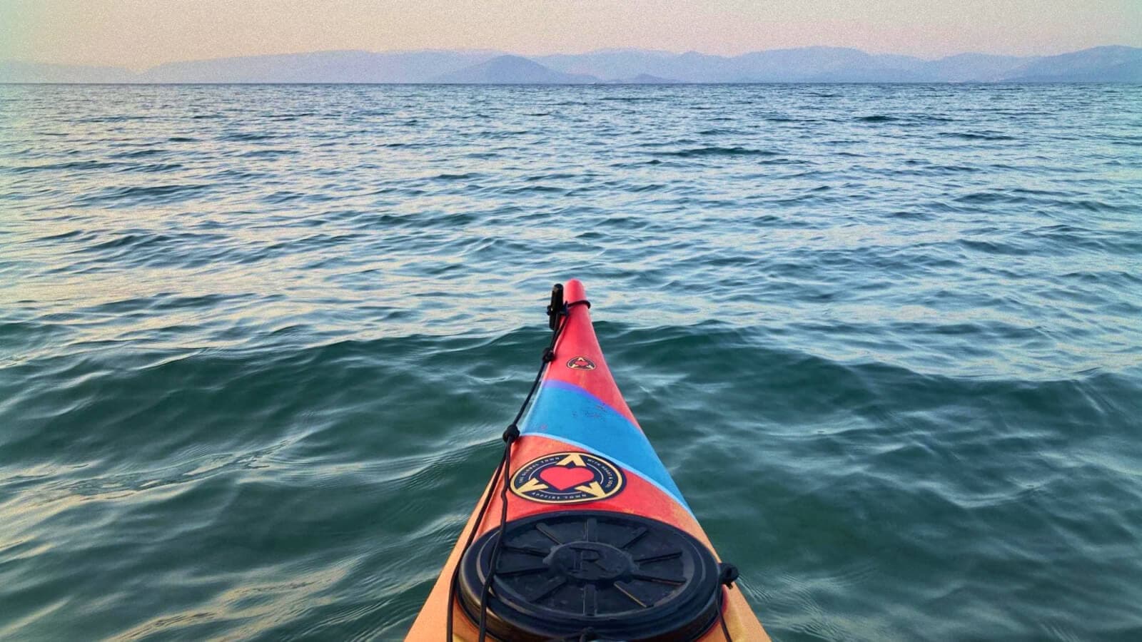 View from the front of a kayak on a calm lake, with a paddle decorated with a logo in the foreground and distant mountains on the horizon.