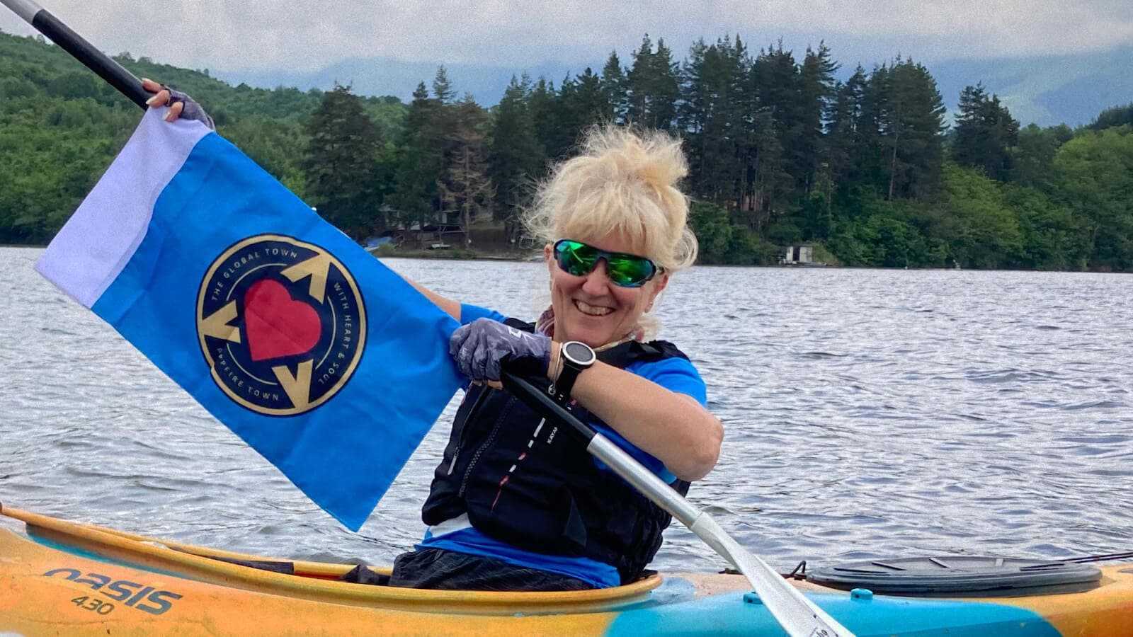 Woman kayaking on a lake, smiling and holding up a paddle decorated with a logo, with trees and mountains in the background.