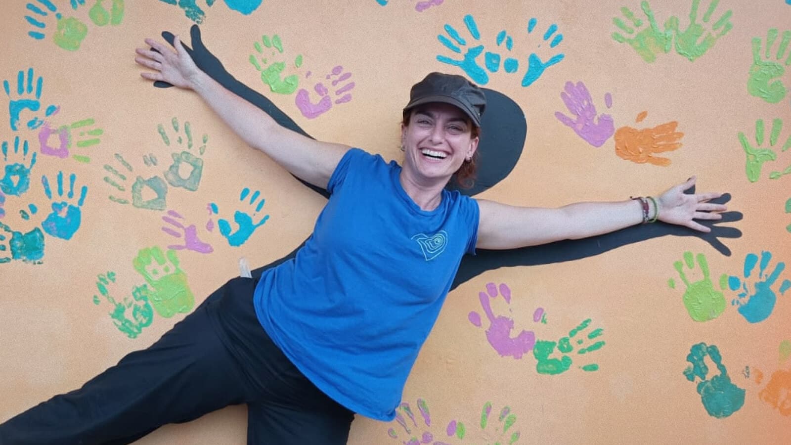 Smiling woman in a blue shirt and black cap posing playfully against a wall painted with colorful handprints, arms stretched wide in front of a black silhouette outline.