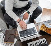 Overhead view of a person working on a laptop near fast food, notebook, and MIDI keyboard in a casual workspace.