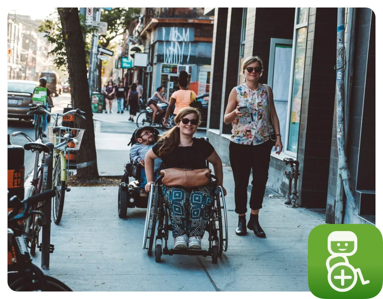 Two women, one in a wheelchair, and a man in a mobility device smiling and moving along a city sidewalk lined with bicycles, with pedestrians and storefronts in the background.